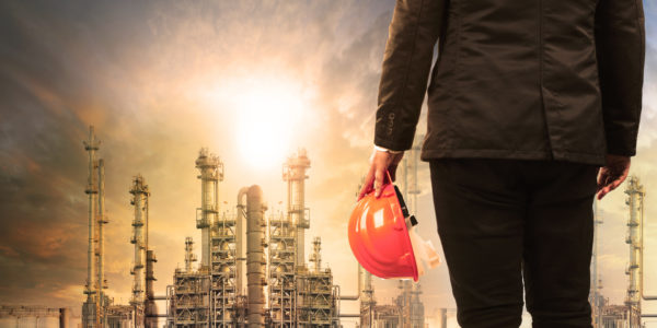 engineering man with safety helmet standing in industry estate against sun rising above oil refinery plant